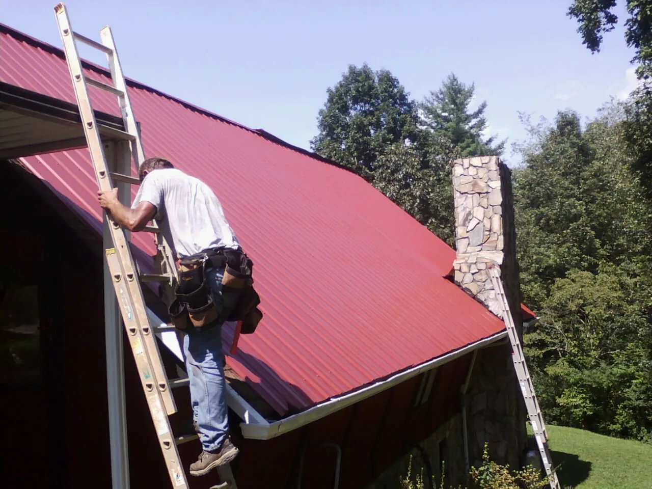Roofer on a ladder beside a red metal roof and stone chimney — Soffit & Fascia Repair by Roofing Brothers in Springville